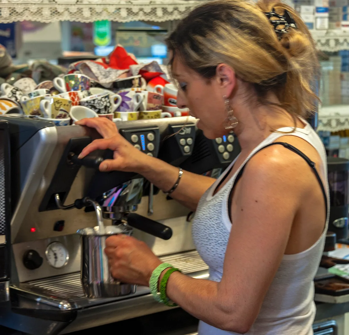 Barista donna di profilo usa macchina espresso professionale; sullo sfondo decine di tazzine colorate impilate in modo caotico.