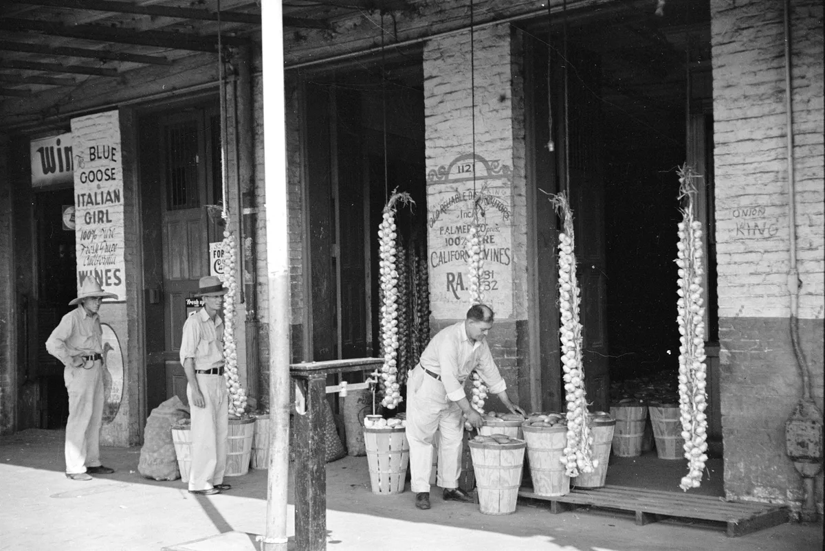 Foto anni '40 di un mercato americano: tre uomini tra ceste di verdure e filze di cipolle appese, insegne dipinte sul muro.