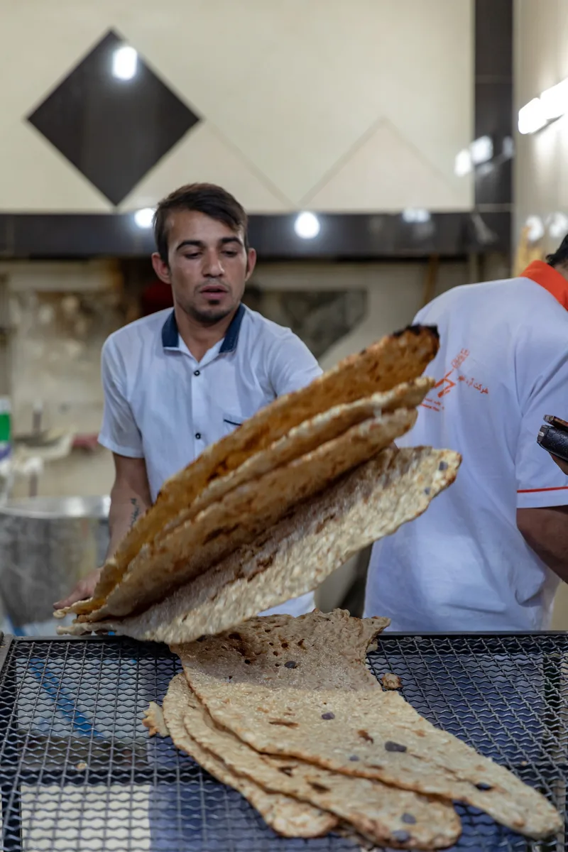 Fornaio in maglietta bianca che solleva pile di pane lavash appena sfornato su una griglia metallica in una panetteria
