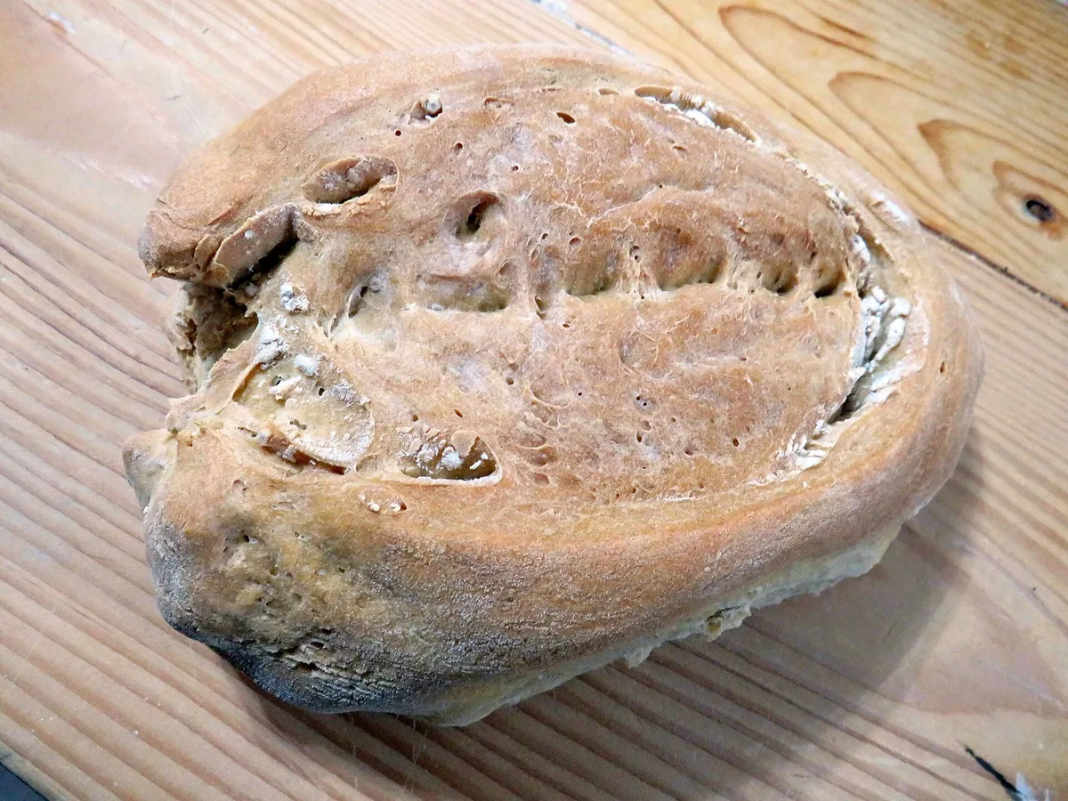 Pagnotta di pane artigianale con crosta irregolare e buchi naturali, appoggiata su un tagliere di legno chiaro.