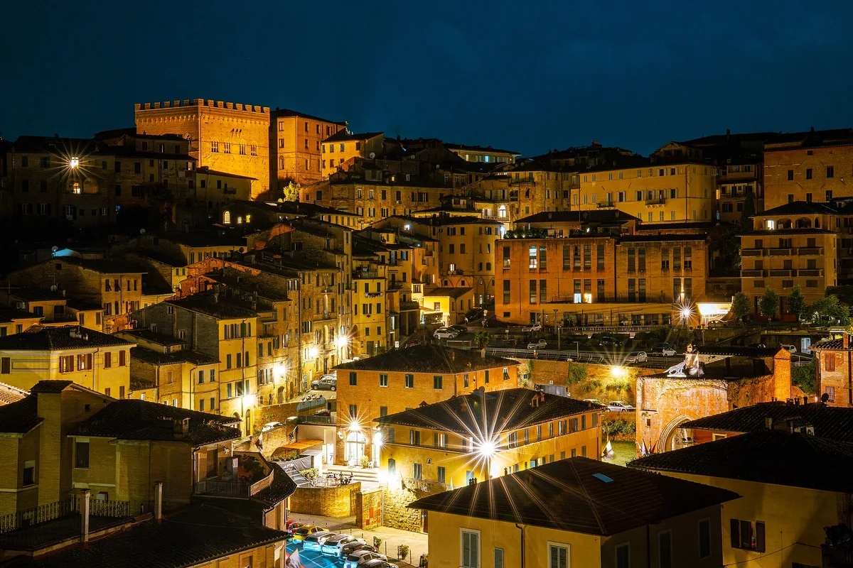 Panorama notturno di Siena con edifici medievali illuminati, luci starburst e torre merlata sullo sfondo blu scuro.
